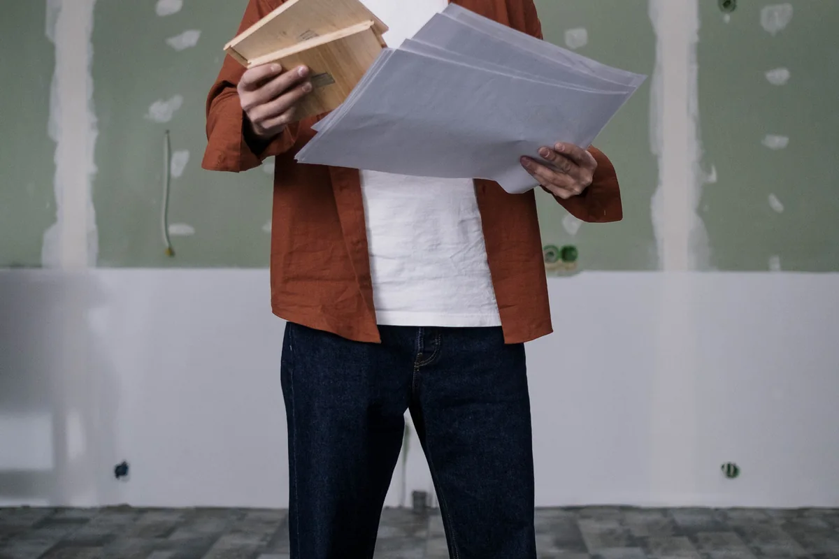 Person comparing wood flooring samples with printed plans in an unfinished room